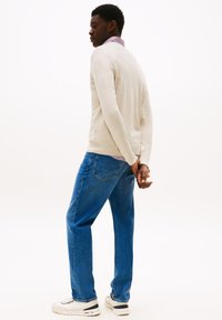 Jeune homme se tenant avec les mains croisées derrière le dos, portant un pull beige, un jean bleu et des baskets blanches sur fond blanc.