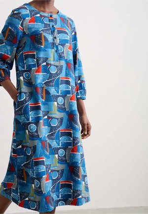 Frau trägt ein blaues Midikleid mit buntem abstraktem geometrischem Muster und Dreiviertelärmeln, steht mit einer Hand in der Tasche.