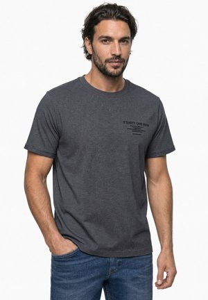 Mand med kort mørkt hår og skæg iført mørkegrå kortærmet skjorte med print og blå jeans, stående med den ene hånd i lommen.