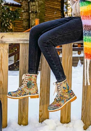 Colorful, patterned lace-up boots with a mosaic design; canvas upper, rubber sole, and white laces, worn with dark jeans, set on a wooden railing.