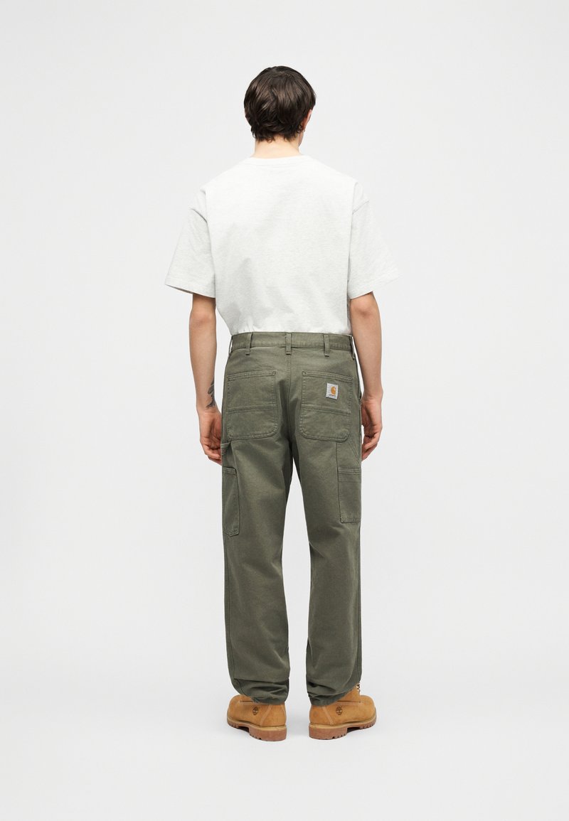 Mann, der olivgrüne Cargo-Hosen, beige Arbeitsstiefel und ein einfaches weißes kurzärmeliges Shirt trägt, steht mit dem Rücken zur Kamera vor einem schlichten Hintergrund.