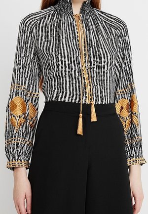 Blouse à motifs noir et blanc avec des formes géométriques brodées en or sur les manches et un lien à glands dorés au col, portée avec un pantalon noir.