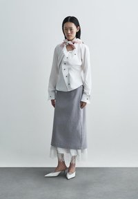 Λευκό πουκάμισο με κουμπιά και ροζ πασμίνα, συνδυασμένο με γκρι φούστα με δαντελένιο τελείωμα. Το μοντέλο φοράει λευκά ψηλοτάκουνα παπούτσια. Μινιμαλιστικό φόντο.