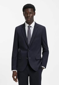 Abito blu navy scuro in tessuto texturizzato con taglio sartoriale, chiusura a due bottoni e rever a lancia. Abbinato a una camicia bianca e una cravatta a fantasia.