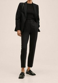 Černé upravené sako s odpovídajícími vypasovanými kalhotami, žebrovaný černý top a lesklé černé mokasíny. Hladká textura a čisté linie.