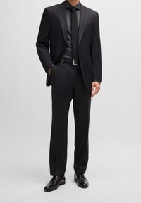 Schwarzer, maßgeschneiderter Anzug mit einreihigem Sakko, passendem Hemd und Krawatte. Die Hose hat einen geraden Beinabschnitt. Schwarze Lederschuhe.