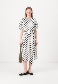 Vestido branco com bolinhas pretas, mangas curtas bufantes, cintura cintuada, silhueta em A, combinado com sapatilhas pretas e uma bolsa de ombro verde oliva.