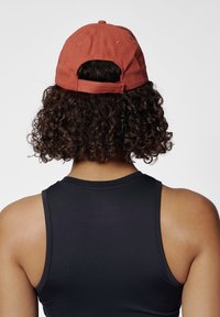 Röd bomullsbasker med böjd skärm, justerbar rem och knappdetalj på toppen, bärs med ett svart ärmlöst linne.