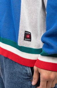 Pulover colorat tricotat, cu dungi albastre, albe și verzi, având un tiv cu dungi roșii și albe. Include un patch cu logo negru, inscripționat cu "KAOTIKO SOCIETY."