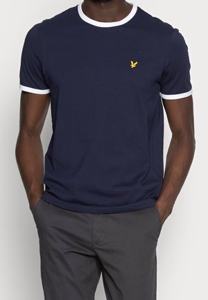 Homme portant un t-shirt bleu marine à manches courtes avec un bord blanc au col et aux manches, associé à un pantalon gris foncé, mains dans les poches.