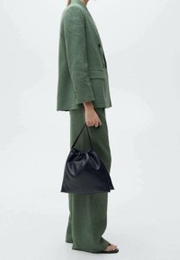 Femme en costume vert olive tenant un sac à main en cuir noir à cordon, debout devant un fond blanc uni.