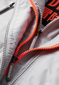 Nærbillede af en grå jakke med to synlige lynlåse, den ene med klare orange tænder, begge mærket med "Superdry" på sorte lynlåstræk.