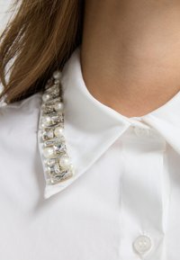 Witte kraagshirt met een decoratieve versiering van parels en rechthoekige kristallen langs de kraag, wat textuur en contrast toevoegt.