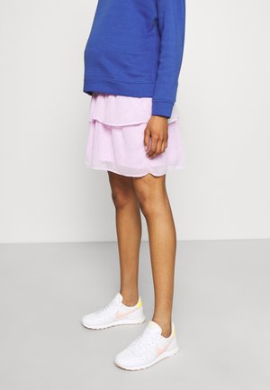 Person iført en blå sweatshirt, lagdelt lyserød nederdel og hvide Nike-sneakers med pink og gule detaljer, stående mod en ensfarvet baggrund.