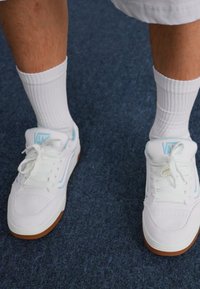 White sneakers with light blue accents, leather upper, perforated toe, round laces, and a gum rubber sole. Worn with white crew socks.