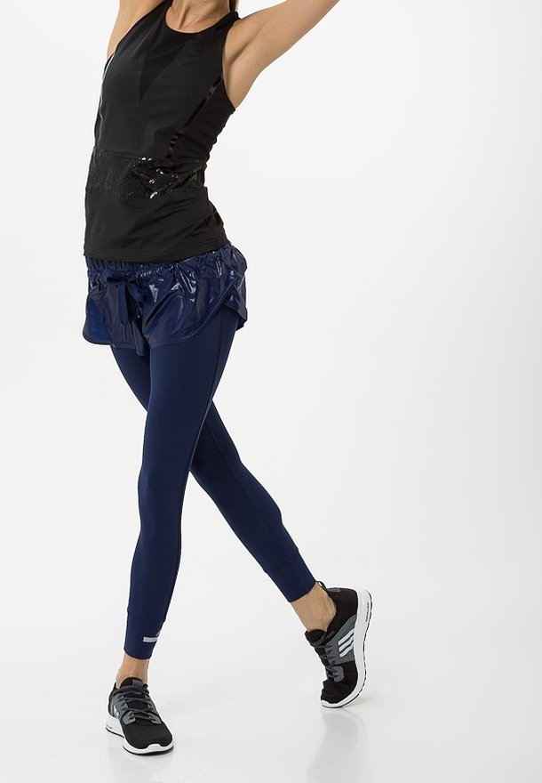 Svart ärmlös topp med glänsande accenter, kombinerad med en blå, glänsande lagerkjol och marinblå leggingar. Svarta träningsskor med vita sulor.