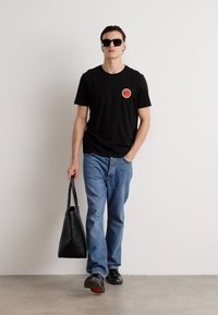 Black T-shirt with a circular red logo, blue jeans, black shoes, and a black tote bag. Subject wears dark sunglasses. Plain background.