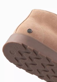 Botas de tobillo de ante en beige con una suela de goma texturizada que presenta un patrón de zigzag, acentuadas con un disco de logo metálico en el lateral.