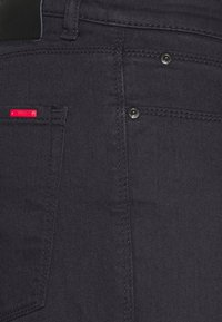 Jeans de denim oscuro con un ajuste slim, costuras visibles, un botón negro y una etiqueta de marca roja en el bolsillo delantero, con una textura suave.