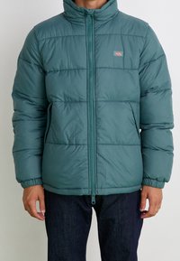 Puffjacka i teal, med hög krage, elastiska muddar och två framfickor. Designen inkluderar horisontell quiltning och zip-stängning.