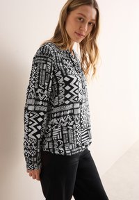 Blouse à motifs noir et blanc avec des designs géométriques, patte de boutonnage et manches longues. Portée avec un pantalon noir sur un fond clair.
