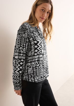 Blouse à motifs noir et blanc avec des designs géométriques, patte de boutonnage et manches longues. Portée avec un pantalon noir sur un fond clair.
