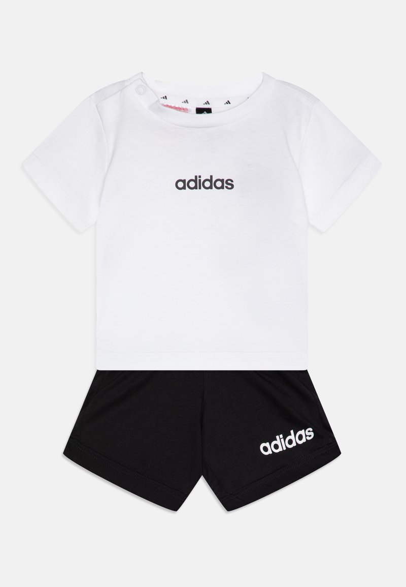 Bijela pamučna majica s crnim "adidas" logotipom, kratkim rukavima i dugmetom na ramenu, u kombinaciji s crnim kratkim hlačama koje imaju bijeli logotip.