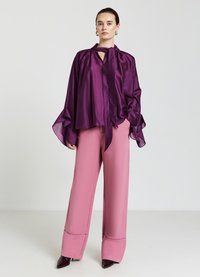 Frau steht da und trägt eine schimmernde lila Bluse mit einer Halstiefe, weit geschnittenen Ärmeln, kombiniert mit hoch taillierten, pinken, weiten Hosen und dunklen Stiefeln.