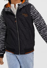 Giacca nera trapuntata con maniche a motivo zebra, dettagli arancioni e zip frontale. Presenta un cappuccio e polsini e orlo a costine.