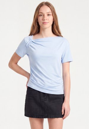 Jeune femme aux longs cheveux châtain clair portant un haut bleu clair à manches courtes avec un détail noué et une jupe en jean noire, debout avec une main sur la hanche.