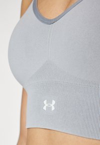 Sujetador deportivo en gris claro con textura acanalada. Presenta un escote redondeado y un pequeño parche de logo blanco. Diseño sin costuras para mayor comodidad.