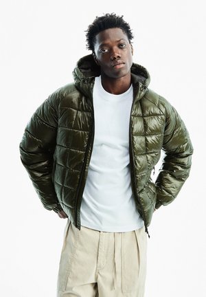 Jeune homme aux cheveux courts et bouclés portant une doudoune vert foncé, un t-shirt blanc et un pantalon beige, debout les mains sur les hanches devant un fond blanc.