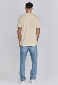 Beige T-Shirt mit kurzen Ärmeln, aus Baumwolle, Rundhalsausschnitt, lässige Passform, kombiniert mit hellblauen Jeans, getragen mit weißen Sneakern.