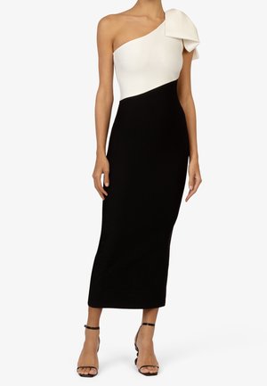 Kobieta w czarno-białej sukience na jedno ramię z dużą kokardą na ramieniu, w połączeniu z czarnymi szpilkami na kostkę.