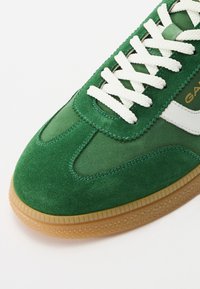 Adidas verde din suede cu șireturi albe și accent lateral; dispune de o talpă texturată din cauciuc și detalii cusute lângă vârf.