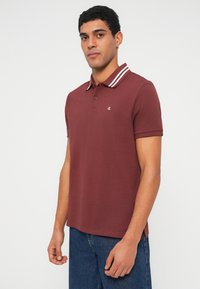 Férfi bordó pólóingben, fehér csíkos gallérral és kék farmerral, egyszínű világos háttér előtt állva.