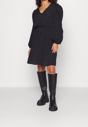 Woman wearing a black long-sleeve dress with puffed sleeves and black knee-high chunky boots, standing against a plain white background.
