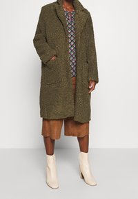 Oliven grøn, fluffy frakke med krave, to frontlommer og et struktureret stof. Parret med en mønstret top og brune shorts.