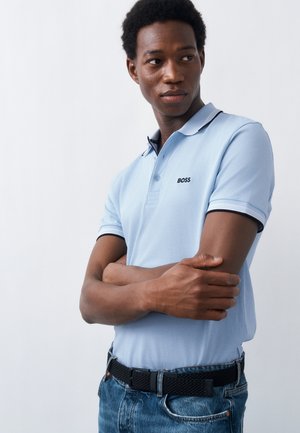 Junger Mann, der ein hellblaues BOSS-Poloshirt und Jeans trägt, steht mit verschränkten Armen da und schaut seitlich vor einem einfarbigen Hintergrund.