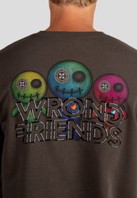 Felpa marrone scuro con tre design di personaggi stilizzati in verde, blu e rosa. La parte anteriore presenta la scritta "WRONG FRIENDS" in lettere grasse e testurizzate.