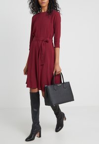 Bordowa sukienka z rękawami 3/4 i wiązaniem w talii, zestawiona z czarnymi botkami sięgającymi za kolano oraz usztywnioną czarną torebką z niebieskimi akcentami.