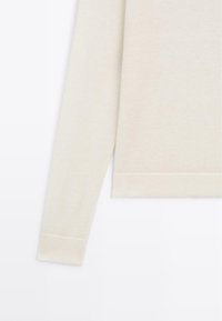 Pull beige au texture lisse, avec un col et des poignets côtelés, et des manches longues. Fini en bas, mettant en avant la douceur du tissu.