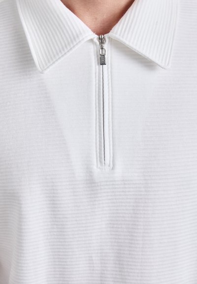 Polo shirt blanche avec une texture côtelée, dotée d'un col zippé. La fermeture éclair a un tirette en argent, et le tissu semble doux et léger.