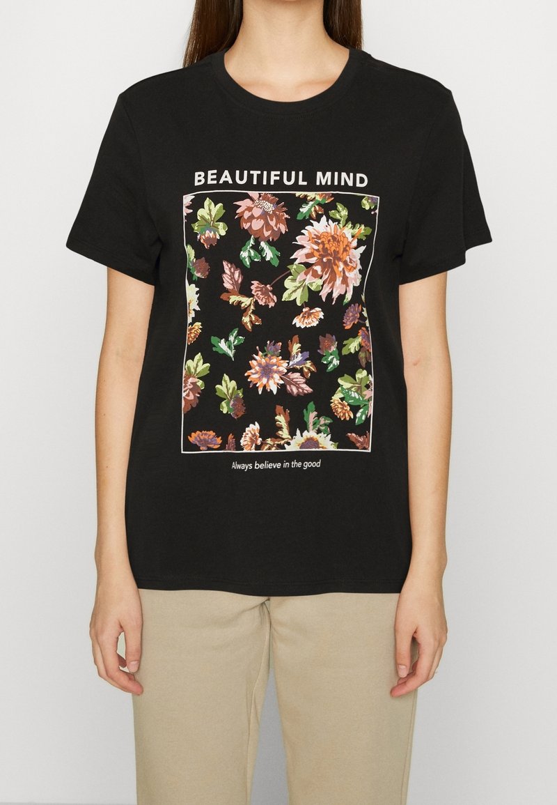 Femme portant un t-shirt noir avec un motif floral et le texte "BEAUTIFUL MIND" au-dessus et "Croyez toujours au bien" en dessous des fleurs.