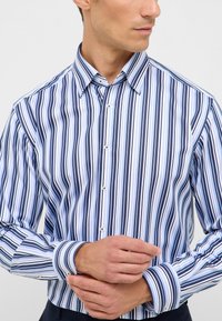 Camicia a righe nei toni del blu, bianco e blu navy; presenta un colletto classico, maniche lunghe e dettagli a contrasto sui polsini.