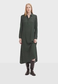 Long-sleeve dress with a green and black diamond pattern, buttoned collar, and asymmetric hem, paired with knee-high brown boots.