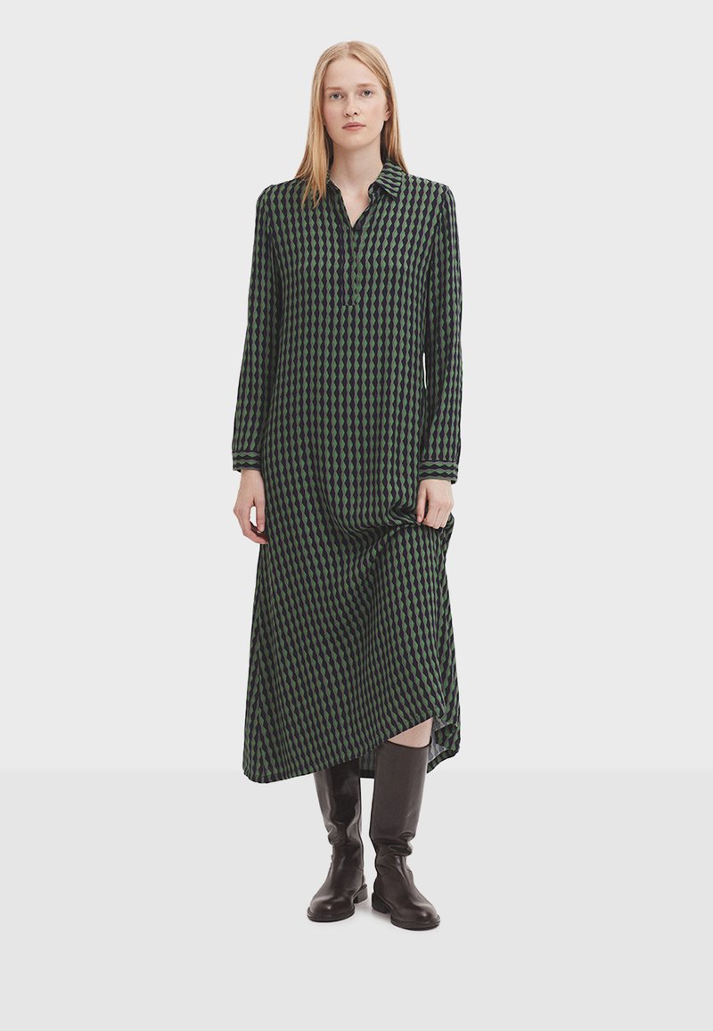Long-sleeve dress with a green and black diamond pattern, buttoned collar, and asymmetric hem, paired with knee-high brown boots.