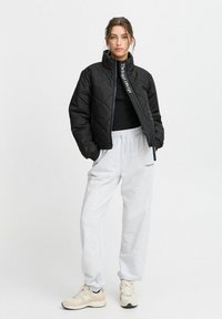 Giacca a vento trapuntata nera con colletto alto, chiusura con zip e dettaglio del logo bianco. Abbinata a pantaloni del tuta grigi e sneakers beige.