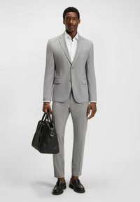 Costume ajusté gris clair avec un pantalon slim et une veste assortie, accompagné d'une chemise blanche et de chaussures en cuir noires, tenant un sac noir.
