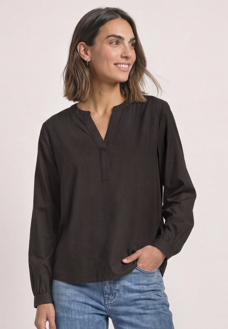 Femme aux cheveux mi-longs portant un chemisier noir à manches longues et un jean bleu, souriante et regardant sur le côté avec une main dans la poche.